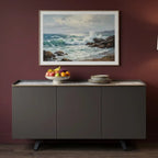 A maroon  wall, with a seascape picture on it and a all windows with beige curtains, and a dark gray sideboard underneath.
