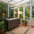 A modern dark gray sideboard cabinet in a bright glass conservatory with terracotta tile floors, a woven area rug, and potted plants.