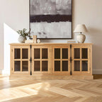 A light natural wood sideboard cabinet with four glass-paneled doors stands against a beige wall in a styled room. Books, vase and a table lamp sit on top, with a large abstract gray wall art piece hanging above.