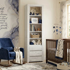 A tall white wooden nursery bookcase with open shelves and two drawers stands between a blue rocking chair and a dark wood crib in a styled nursery room.