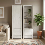 A tall white wooden storage cabinet shown in a styled living room. One door is closed, and the other is open to reveal three shelves with books and small potted plants, alongside a sofa and a potted fiddle-leaf fig on a window sill.