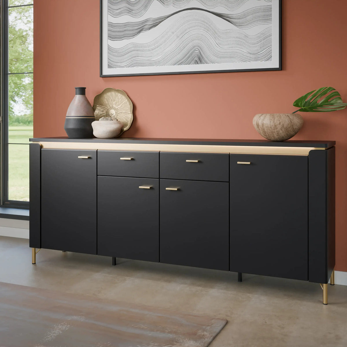 A modern black sideboard cabinet with gold-tone handles and legs, styled in a room with a terracotta wall, decorative vases, and a framed abstract print.