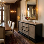 A dark wood and distressed gray sideboard in a traditional dining room, styled with a marble-look top, silver candelabras, a fruit bowl, and a large gold-framed mirror.