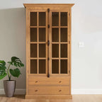 A wooden cabinet with medium brown wood grain, paneled side panel, and glass-front upper doors against a cream wall and pot plant.