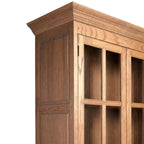 Close-up of a wooden cabinet or bookcase corner with medium brown wood grain, paneled side panel, and glass-front upper doors on a white background.