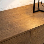 Close-up of a medium brown wooden cabinet or sideboard with visible wood grain, a white wall behind it, and part of a black metal table leg at the top right.