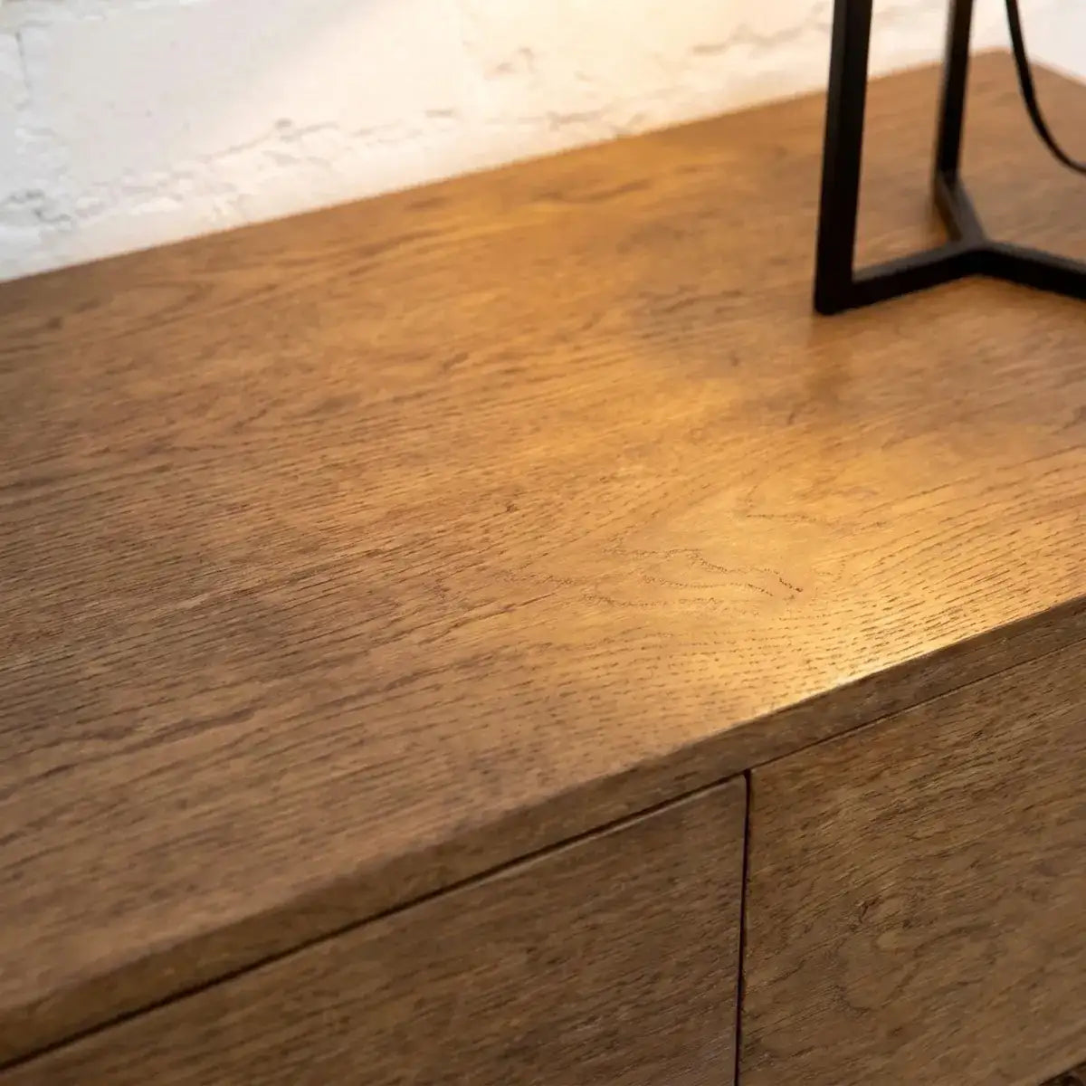 Close-up of a medium brown wooden cabinet or sideboard with visible wood grain, a white wall behind it, and part of a black metal table leg at the top right.