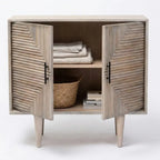 A rustic two-door sideboard cabinet with a weathered gray wood finish, featuring chevron-patterned doors, black metal handles, and tapered legs. The cabinet is shown open with folded linens, a woven basket, and books inside, set against a plain white studio background.