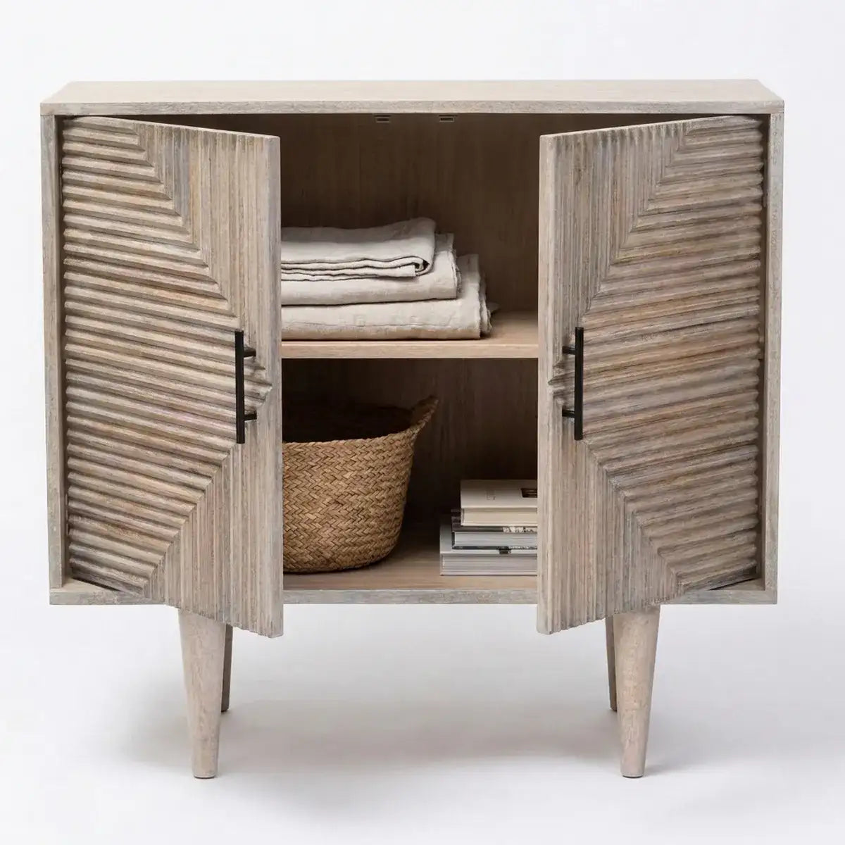 A rustic two-door sideboard cabinet with a weathered gray wood finish, featuring chevron-patterned doors, black metal handles, and tapered legs. The cabinet is shown open with folded linens, a woven basket, and books inside, set against a plain white studio background.