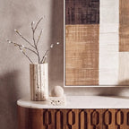 A styled interior scene showing a tall ribbed ceramic vase with small dark speckles, holding bare branches with tiny white blossoms, placed on a curved wooden console table with a white marble top. A large abstract wall art piece hangs above on a textured taupe wall.