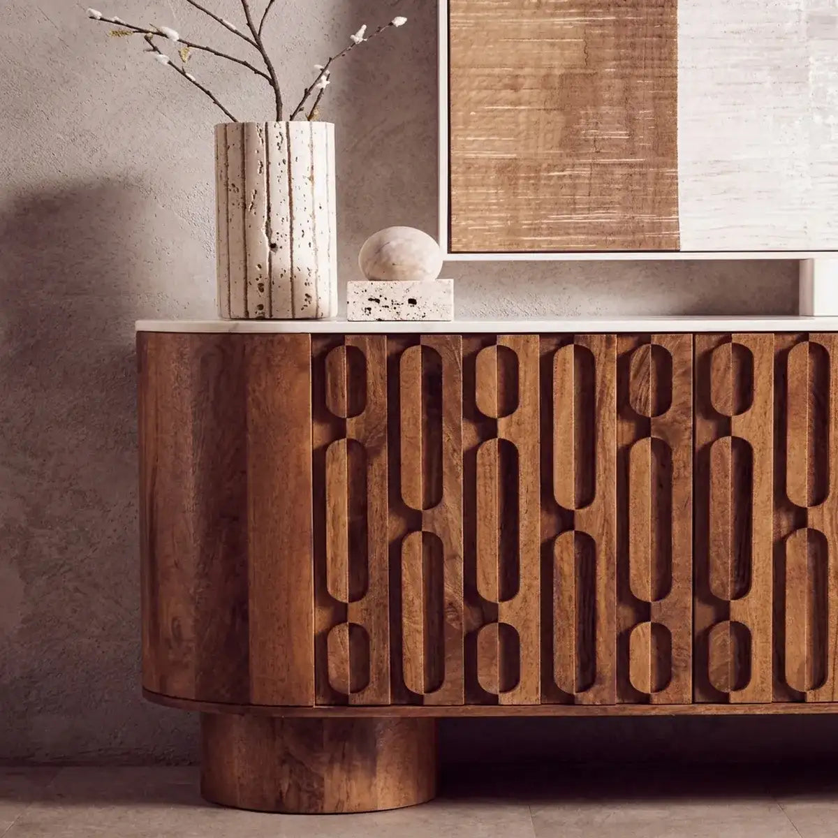 A modern wooden sideboard or credenza with a carved geometric pattern on the front, styled in a minimalist room with a marble top, a textured vase, and wall art.