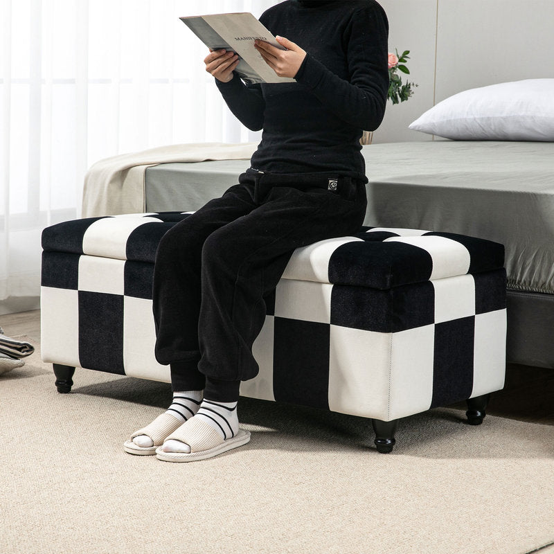 Person sitting on a checkered ottoman reading a book in a bedroom.