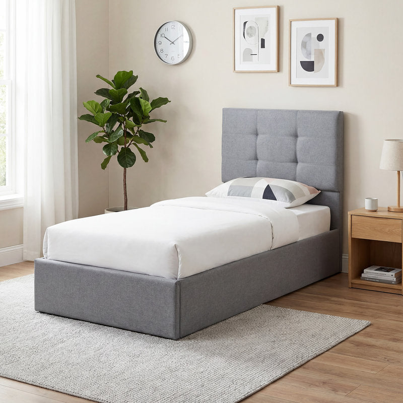 Gray upholstered bed in a bedroom with a plant, clock, and nightstand.