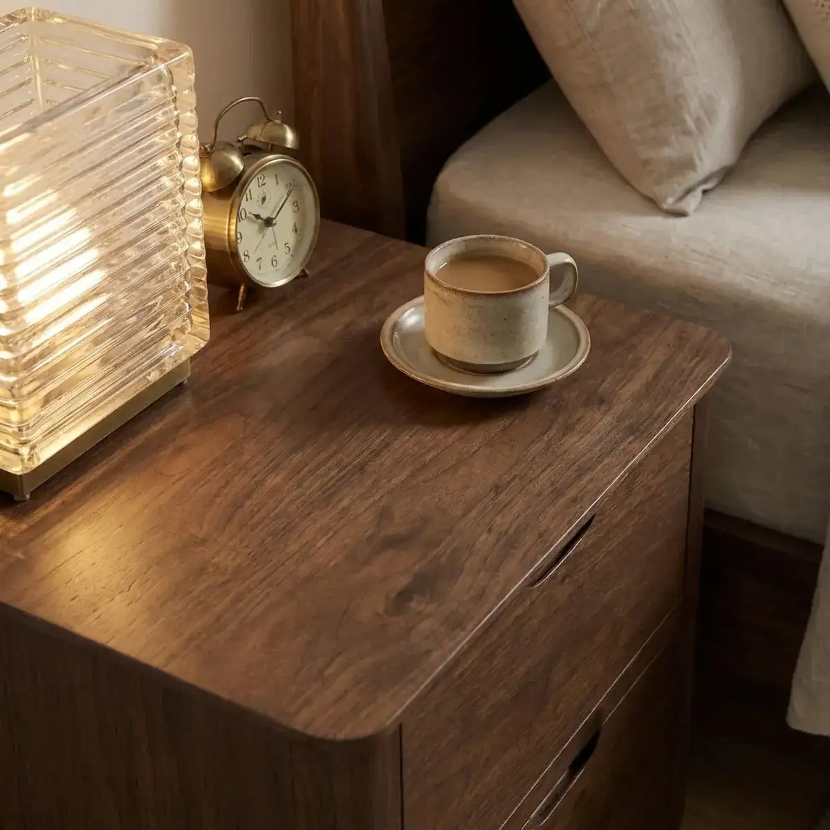 A warm-toned wooden nightstand in a bedroom setting, styled with a glowing ribbed glass lamp, a brass alarm clock, a cup of coffee on a saucer