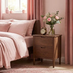 A dark brown wooden two-drawer nightstand in a cozy bedroom setting, styled with a vintage alarm clock and a glass vase of pink and white flowers next to a bed with pink bedding.