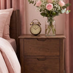 A dark brown wooden nightstand in a bedroom setting, styled with a vintage brass alarm clock and a glass vase of pink roses and baby's breath on top.