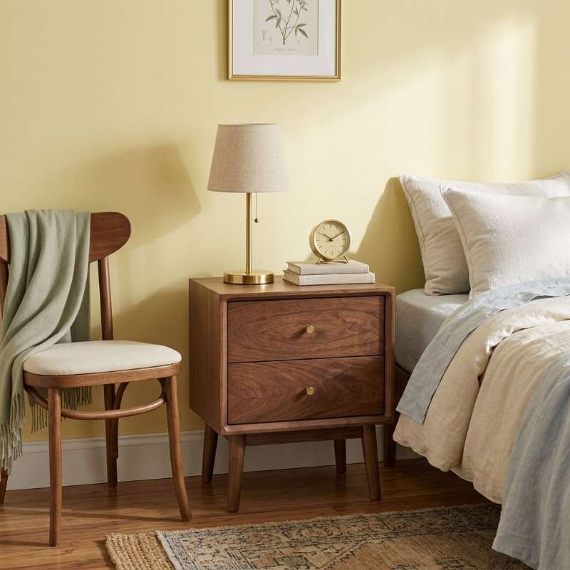 A wooden nightstand with two drawers  styled in a bedroom next to a bed with  a table lamp, a small clock and a chair with a green throw blanket.